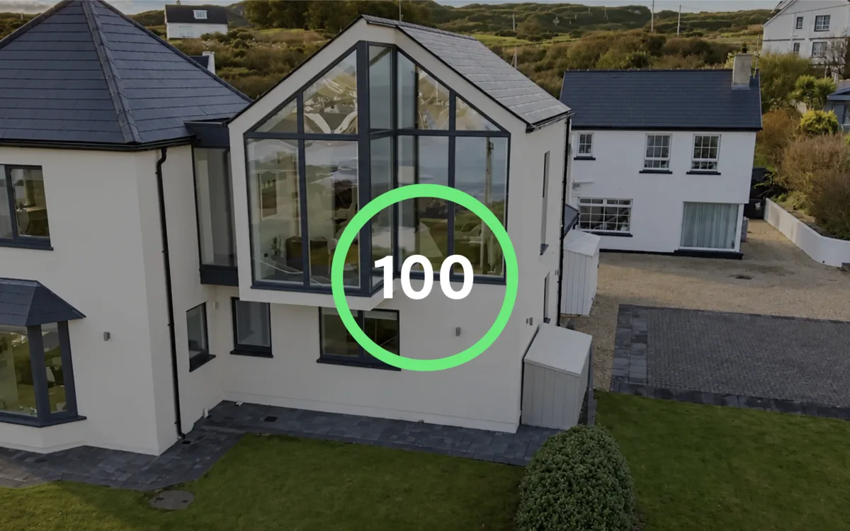 Aerial film still from the Bond Architects homepage, a white rendered house with a glazed gable and a slate roof in Donegal, overlaid with a green Lighthouse score ring reading 100